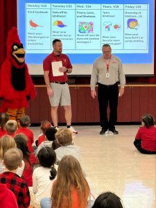 students sitting on gym floor for an assembly with their principal, teacher and mascot, Fireball.