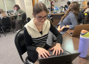 student with brown hair and glasses typing on a computer in a class setting