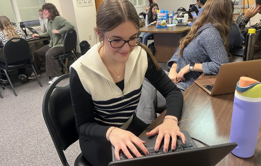 student with brown hair and glasses typing on a computer in a class setting