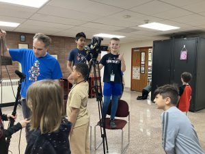 students standing on chairs to see behind video camera and other crew preparing for filming