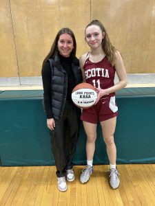 coach stands beside a player in uniform while holding a basketball with 1000 points written on it