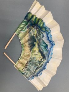 a thick material folded like a fan with sticks on both sides of the material. The pattern starts at the top with white then shades of blue, and greens and browns and yellows.
