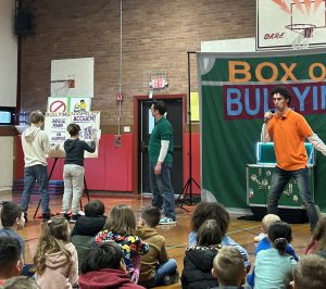 kids moving words around on a whiteboard while one adult watches them and another adult holds a microphone