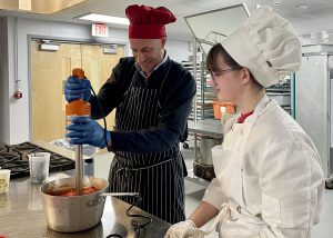 kitchen setting, adult in chefs hat stirring sauce and student in chefs uniform watches