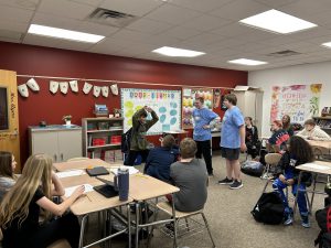 Student wearing backpack stands in front of the class doing jumping jacks as students watch from their seats.